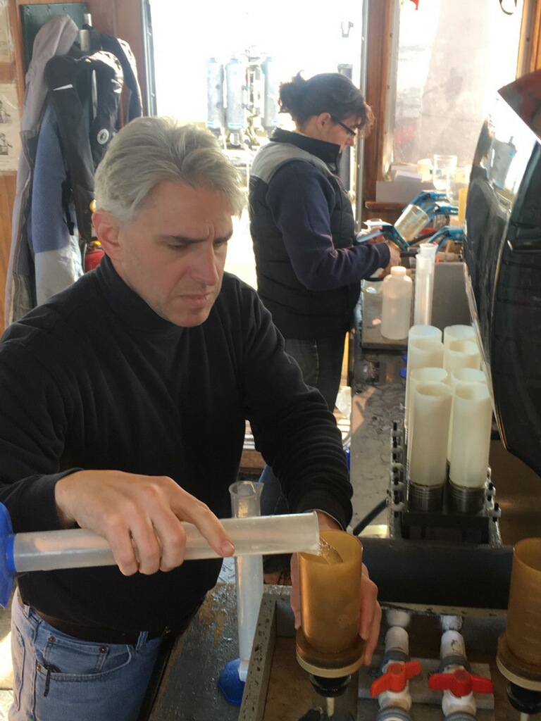 Matt Lyman, an environmental analyst at CT DEEP, pours a water sample into a flask with a filtration device for analysis. In the background, O'Brien-Clayton is preparing to replace filter assemblies into the flasks in order to analyze more samples. Photo credit: CT DEEP.