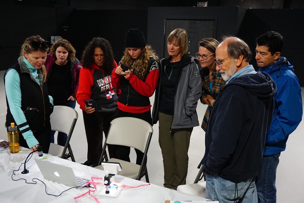 Attendees at a Mentor Teachers Workshop in Sands Point, NY and Mentor Teachers Hildur Palsdottir (third from right) and Leah Master (second from left) huddle at a screen connected to a microscope to view their magnified sample materials from the beach.