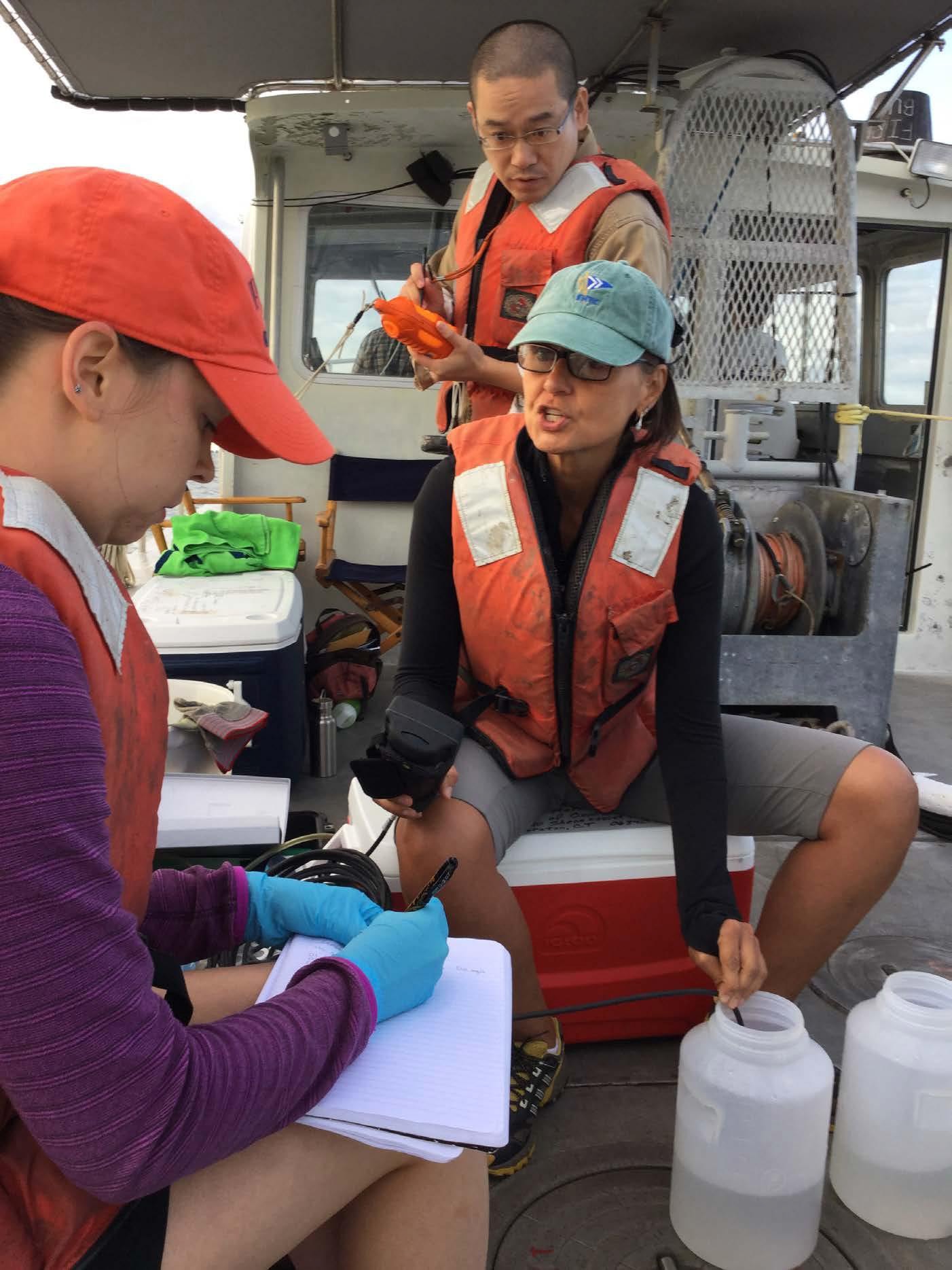 Seated on a boat during a water quality monitoring session,Dr. Penny Vlahos reads data from a multi-probe meter to Allison Byrd. Behind Dr. Vlahos is Yan Jia, a marine sciences graduate student, with a handheld profiler. 