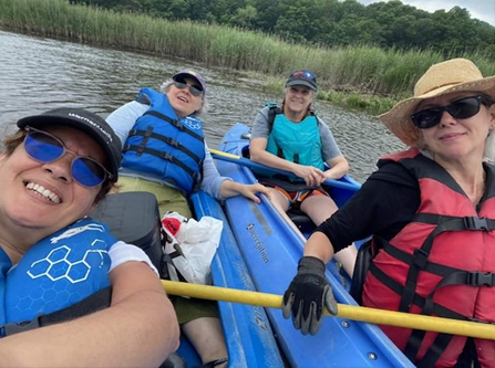A group of four middle-aged women are posed together, with two women in each kayak smiling for the photo. The kayaks are blue and there is green and tan seagrass in the background. The two women in front are wearing sunglasses and hats. They all have life vests, the two women on the left are wearing blue ones. the women on the front right is in read and the back right corner is in a teal vest. 