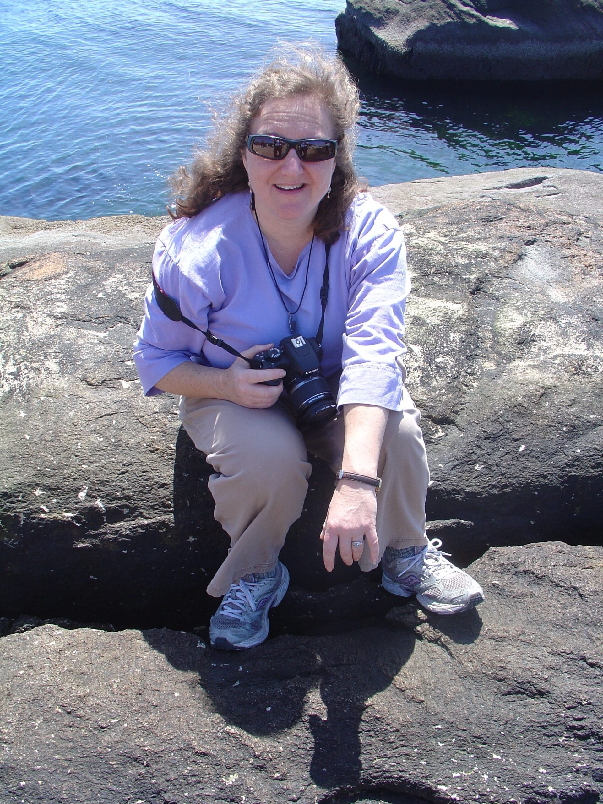Dr. Syma Alexi Ebbin sits on a rock and smiles, holding a camera. 
